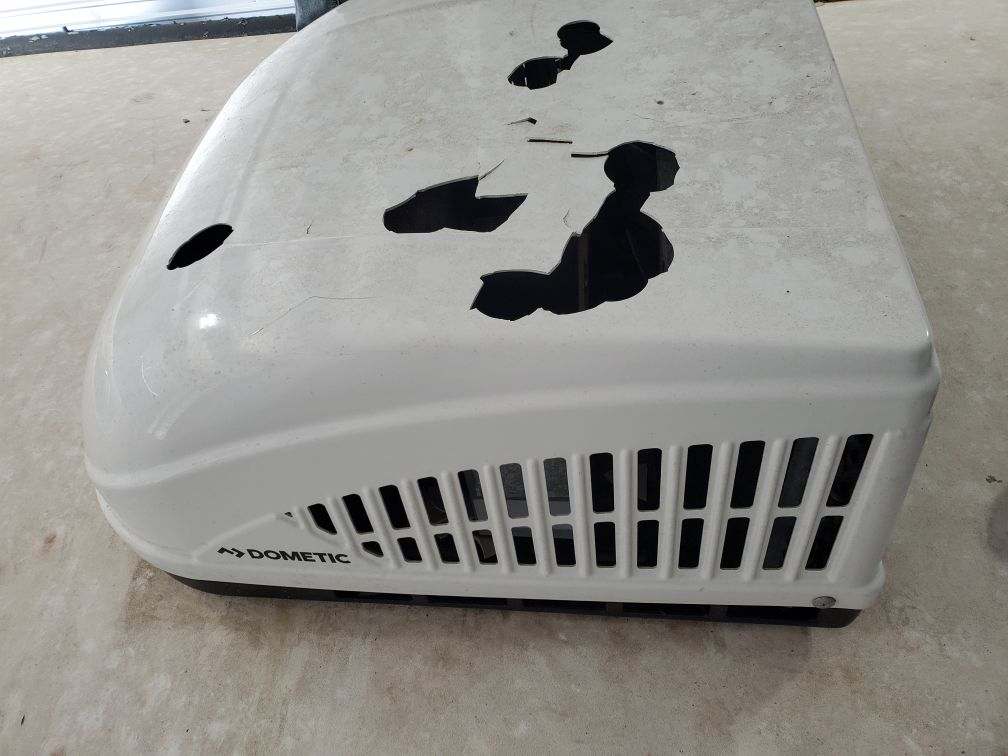 Removed Dometic RV air conditioner shroud on a concrete floor showing catastrophic hail damage — multiple large holes punched through the plastic with jagged broken edges — Boss Bull Mobile RV Services