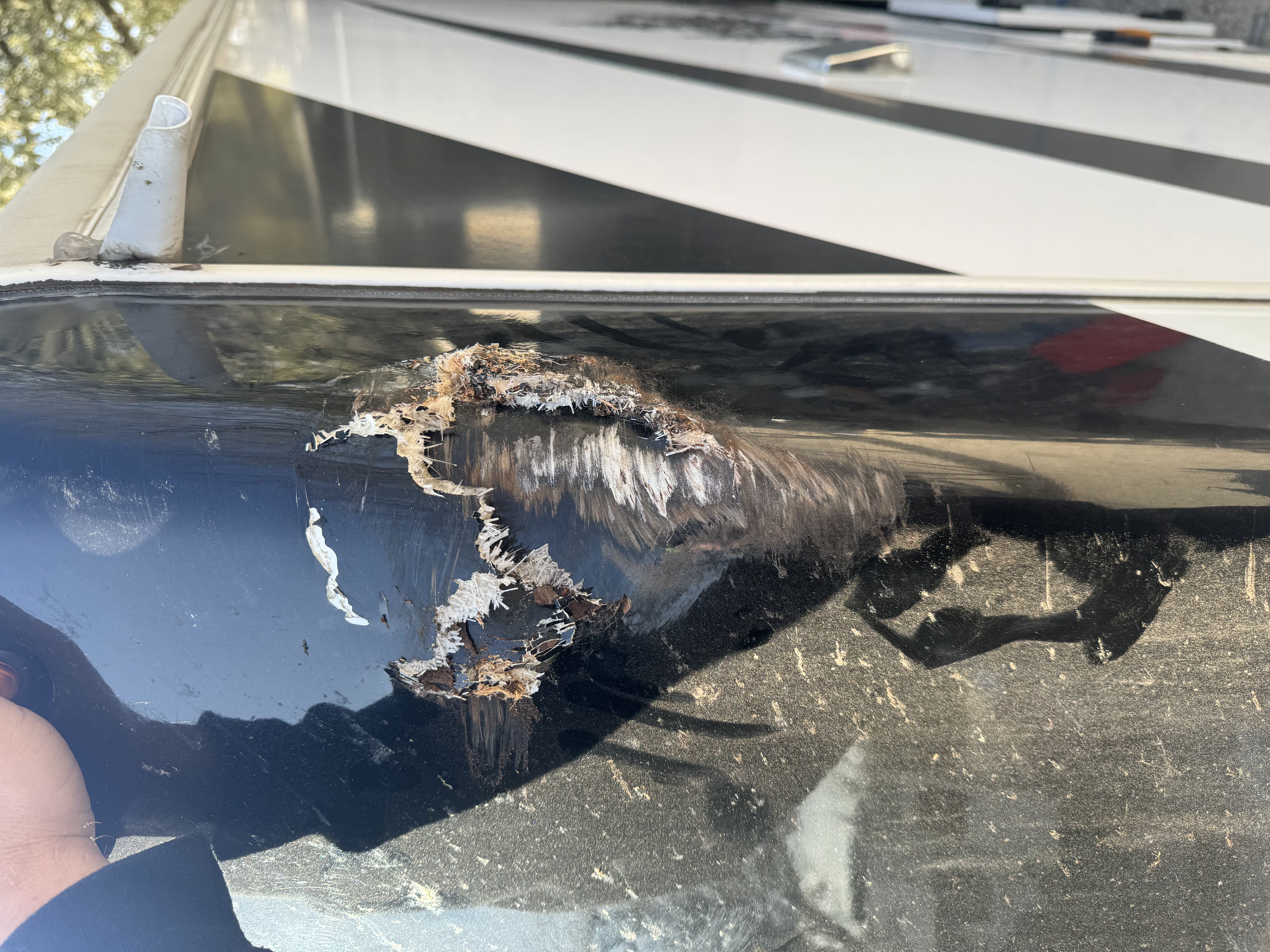 Close-up of a jagged fiberglass tear on an RV front cap — exposed substrate and wood core visible inside the damage crater before insurance claim repair begins — Boss Bull Mobile RV Services