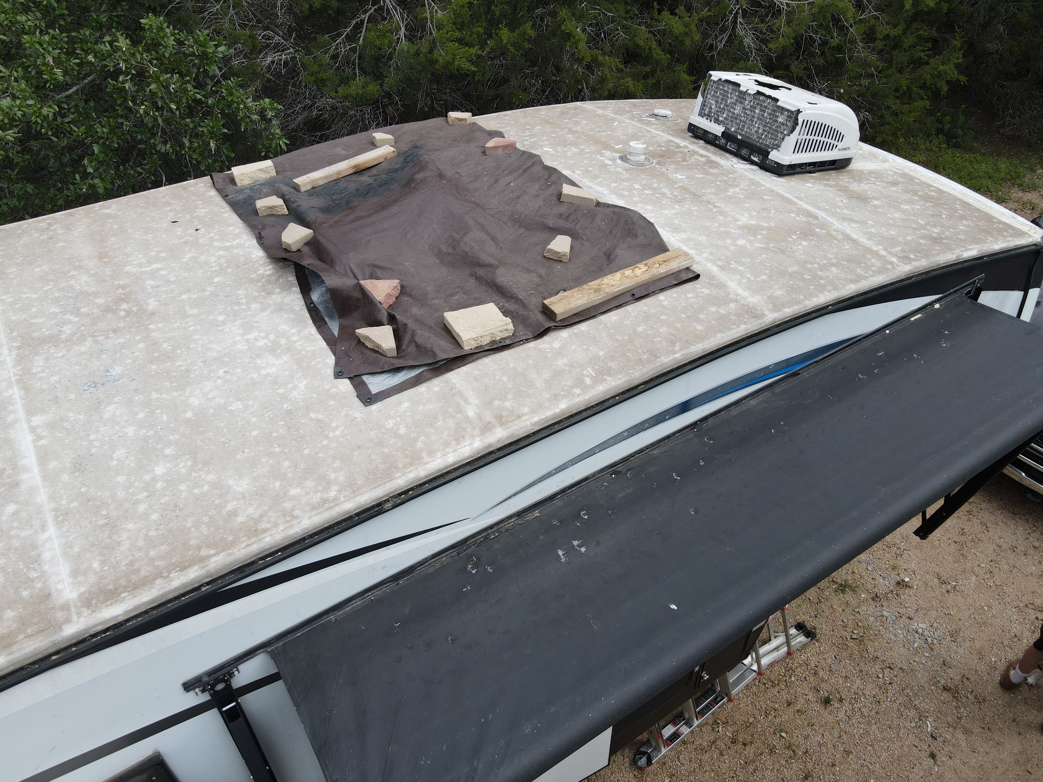 Elevated view of a fifth-wheel RV roof with a large dark brown tarp weighted down by limestone rocks and lumber boards covering a damaged section — emergency field installation — Boss Bull Mobile RV Services