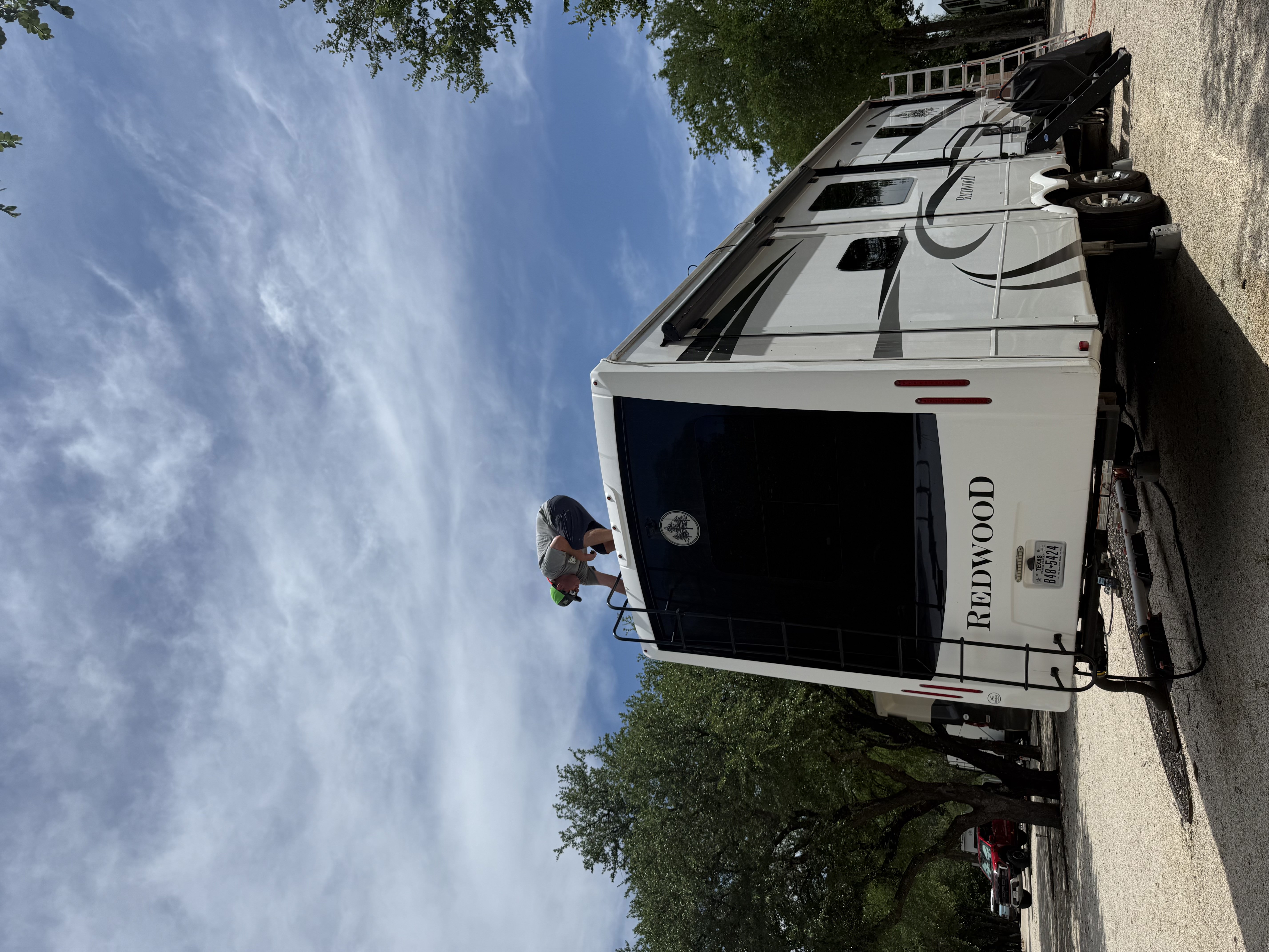 Wide establishing shot of a Crossroads Redwood fifth-wheel at an RV park — Boss Bull technician crouched on the front cap roofline performing roof work, second ladder visible at the rear — mobile on-site roof service — Boss Bull Mobile RV Services