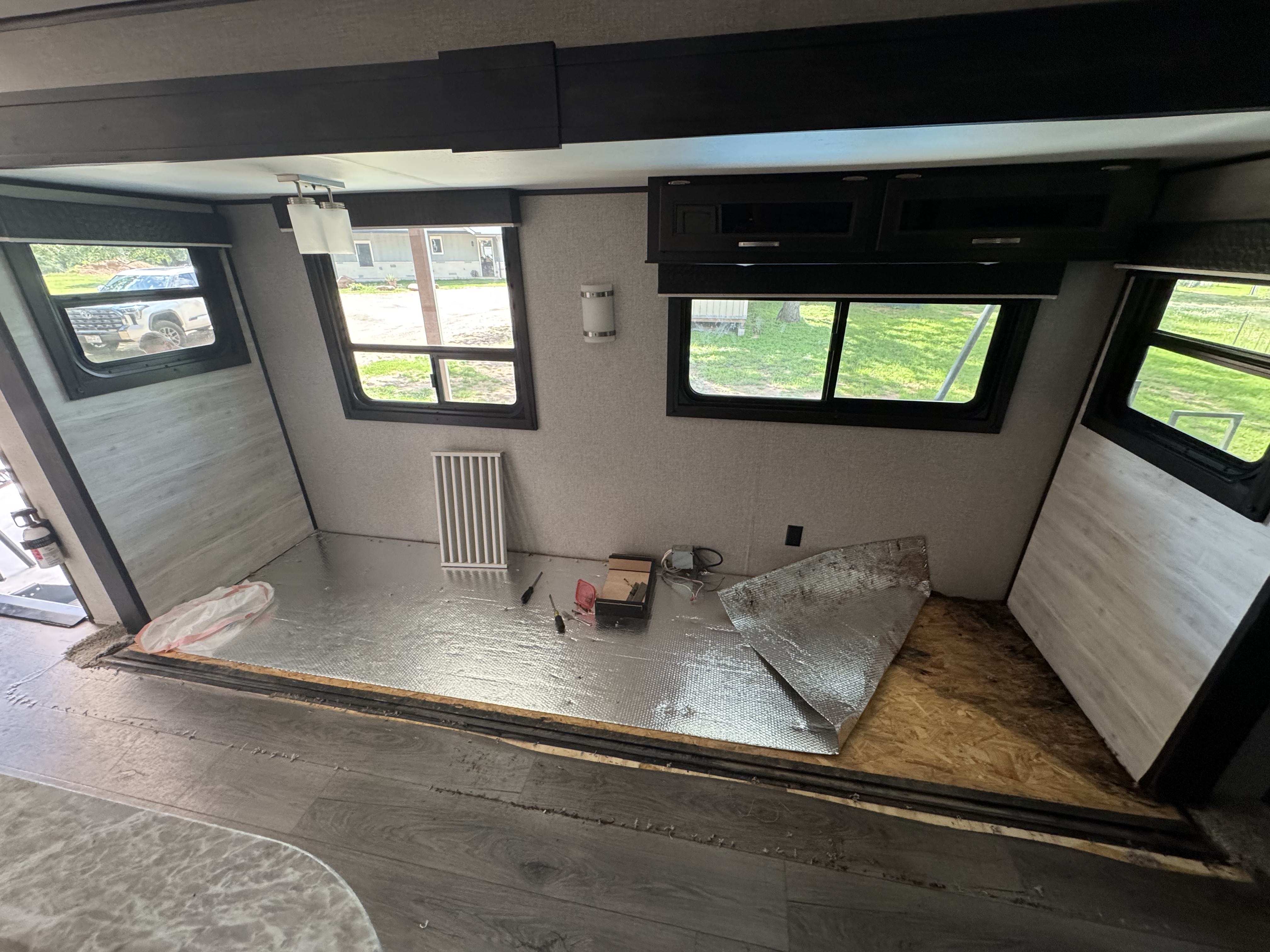 Interior view looking into a stripped RV slide room from the main cabin floor — OSB subfloor exposed in the corner, foil-faced insulation pulled back in the center, floor covering and perimeter trim removed — slide floor replacement in progress — Boss Bull Mobile RV Services