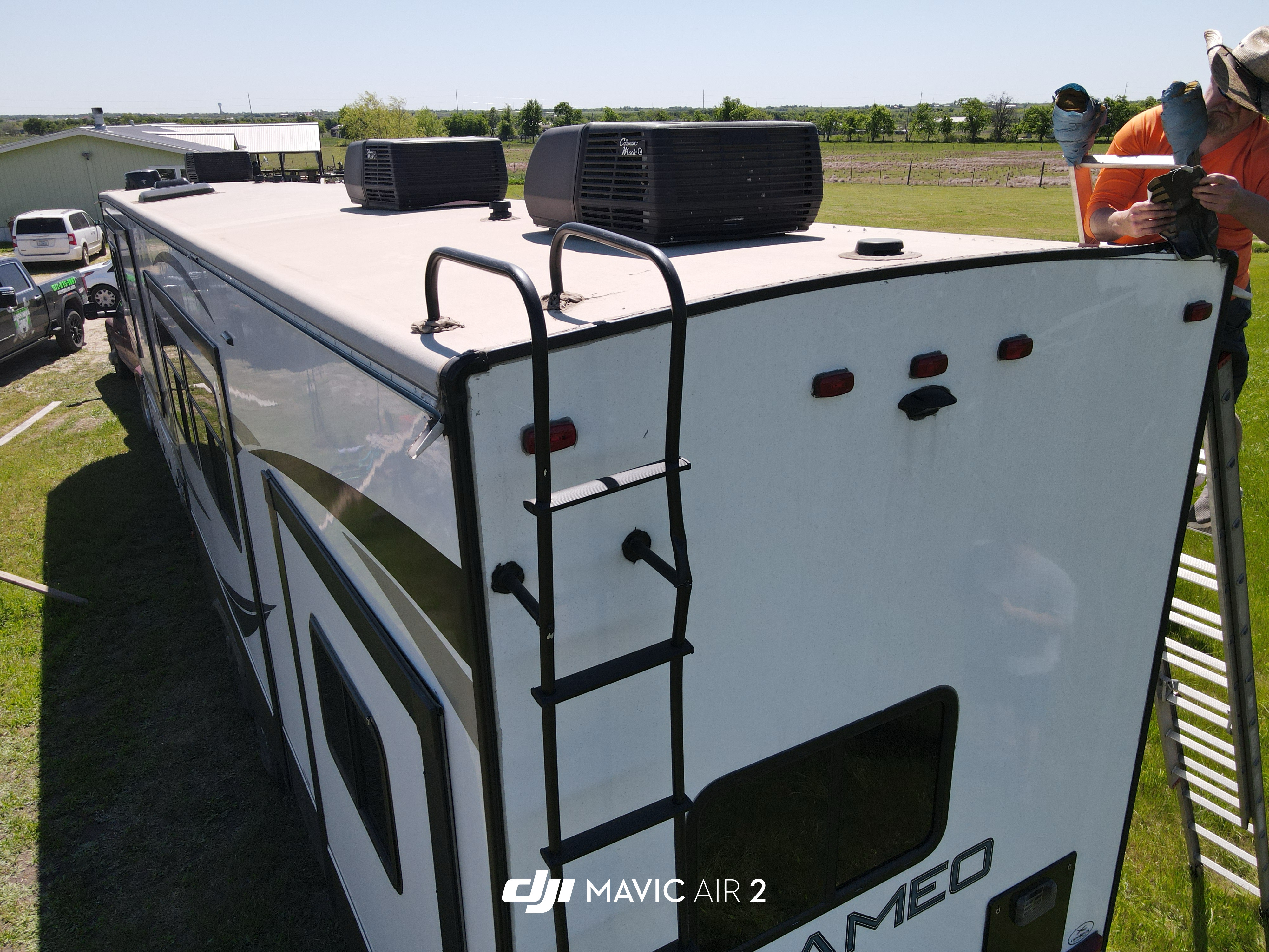 DJI drone shot looking straight down at the rear corner of a Crossroads Cameo fifth-wheel — Boss Bull technician in orange shirt and straw hat fully visible on a ladder with both hands working the roof-to-rear-cap seam on the top face of the unit — Boss Bull Mobile RV Services