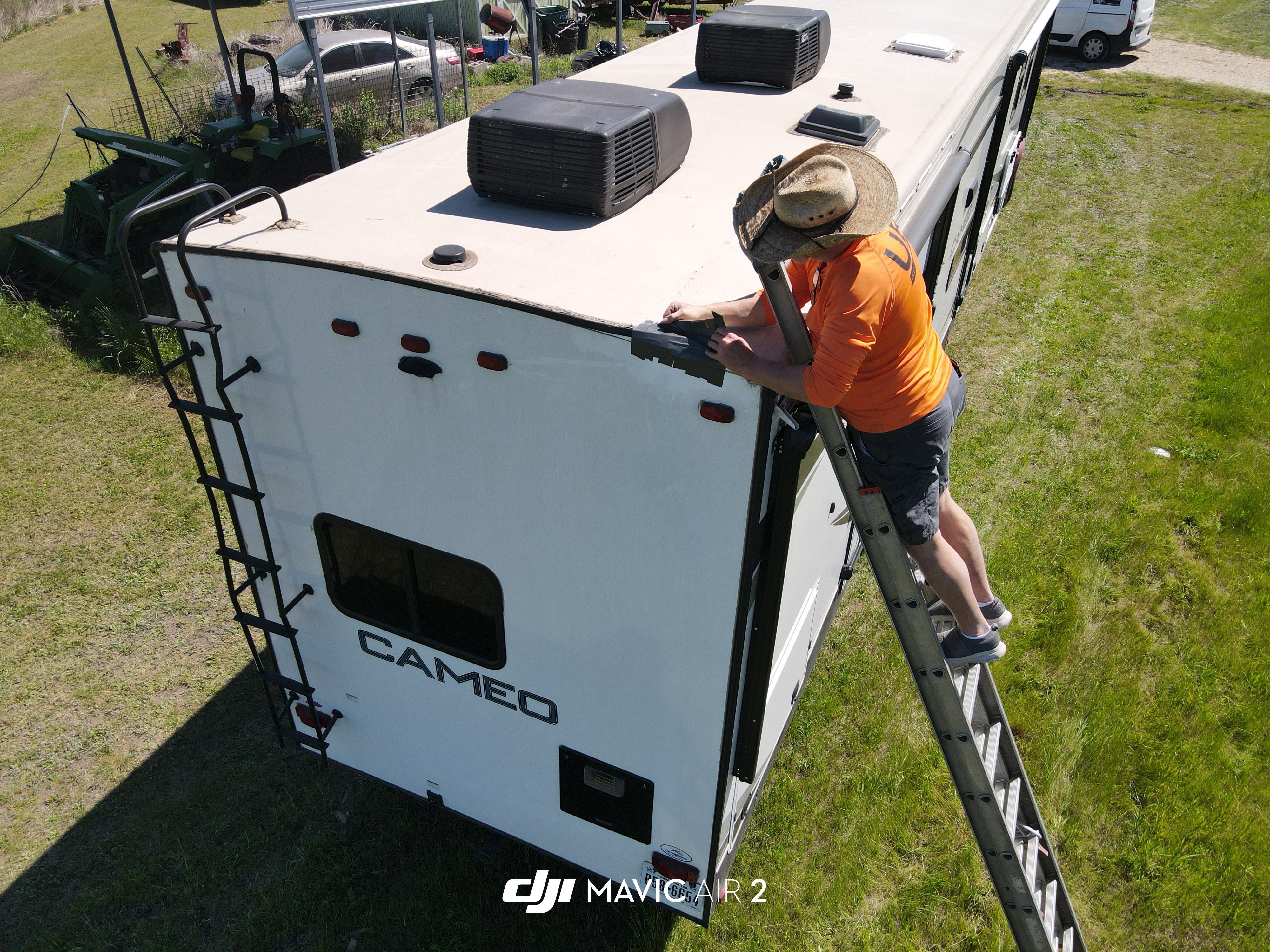DJI drone side-angle shot of a Boss Bull technician in an orange shirt on a ladder applying sealant to the roof-to-rear-cap seam at the top edge of a Crossroads Cameo fifth-wheel — rural Central Texas job site — Boss Bull Mobile RV Services